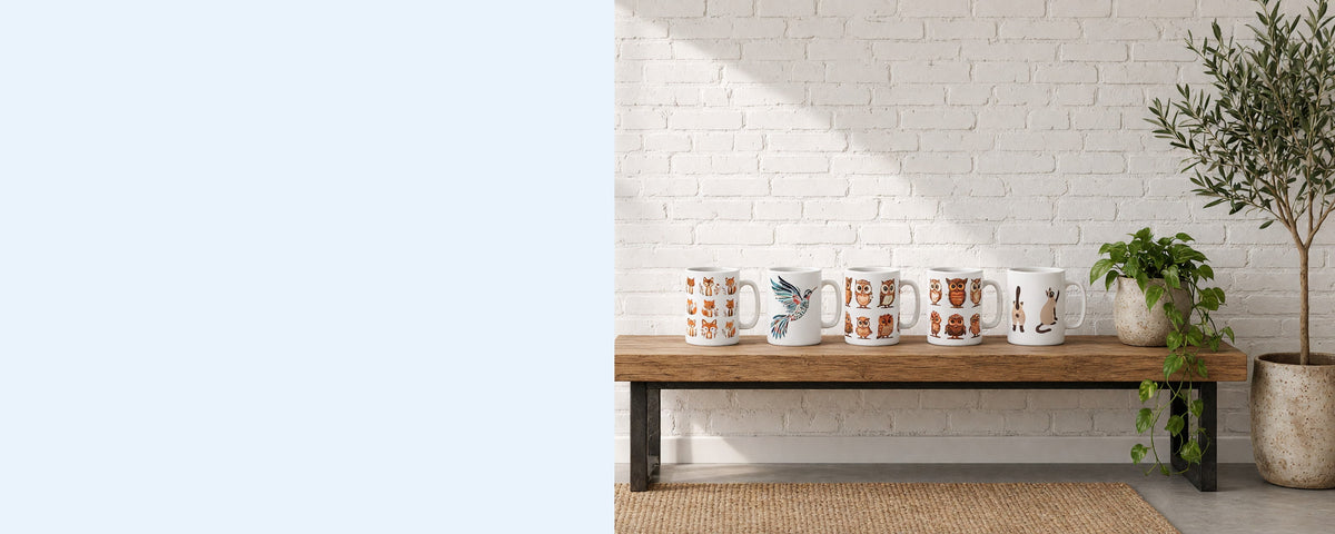 Four printed ceramic coffee mugs in a row on a wooden console table against a sunlit white brick wall, with a pothos plant, an olive branch in a vase and an olive tree