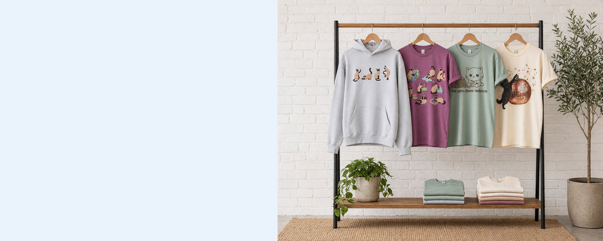 4 printed garments on wooden hangers on a wood and iron clothing rack against a sunlit white brick wall, with an olive tree, trailing pothos and folded tees on the shelf below