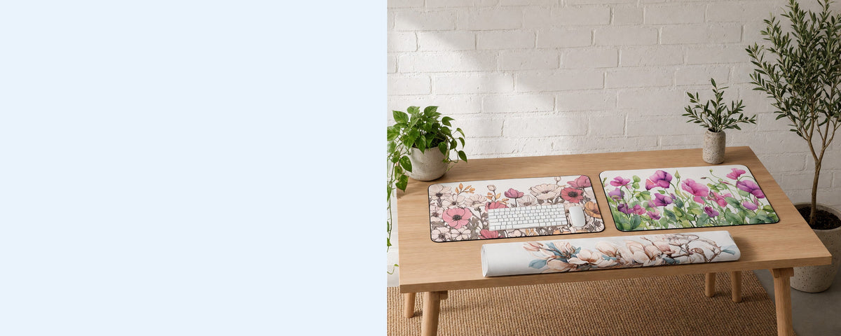 Three printed neoprene desk mats on a tidy desk against a sunlit white brick wall, one in use under a keyboard, one lying flat beside it and one loosely rolled on the desk, with an olive tree