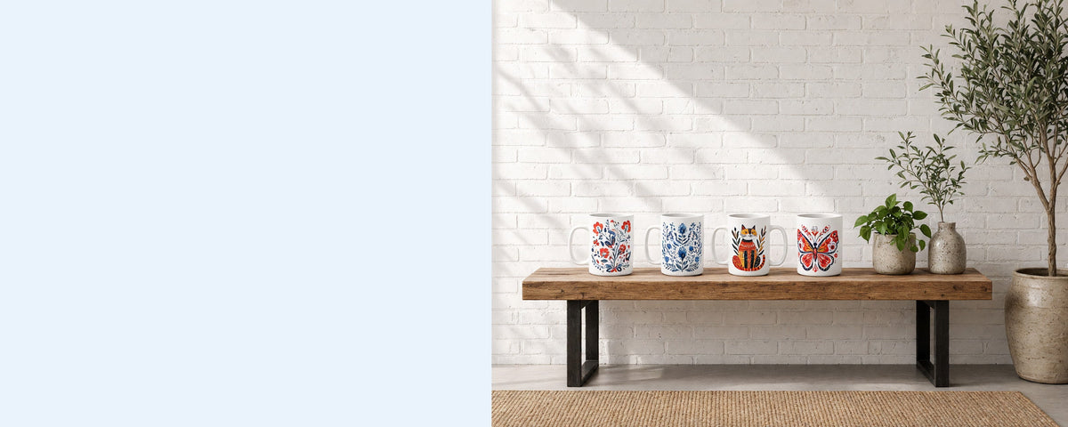 Four printed ceramic coffee mugs in a row on a wooden console table against a sunlit white brick wall, with a pothos plant, an olive branch in a vase and an olive tree