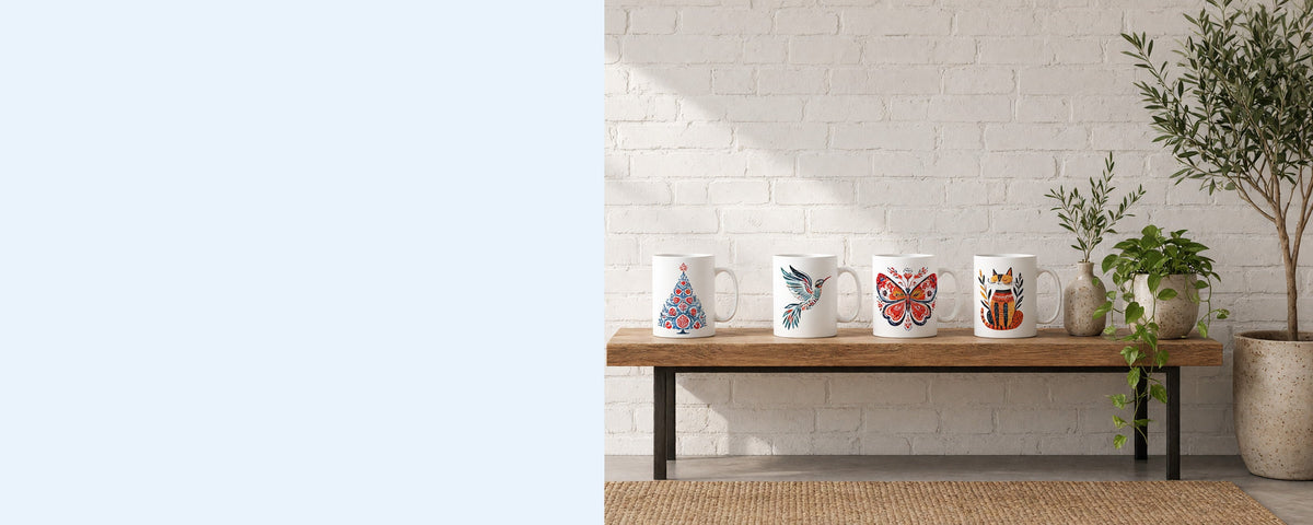 Four printed ceramic coffee mugs in a row on a wooden console table against a sunlit white brick wall, with a pothos plant, an olive branch in a vase and an olive tree