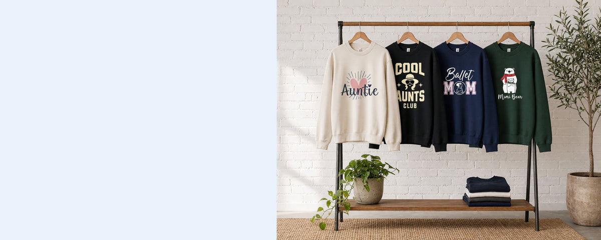 4 printed crewneck sweatshirts on wooden hangers on a wood and iron clothing rack against a sunlit white brick wall, with an olive tree, trailing pothos and folded tees on the shelf below