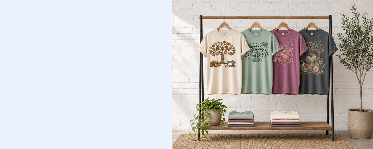 4 printed garment-dyed cotton t-shirts on wooden hangers on a wood and iron clothing rack against a sunlit white brick wall, with an olive tree, trailing pothos and folded tees on the shelf below