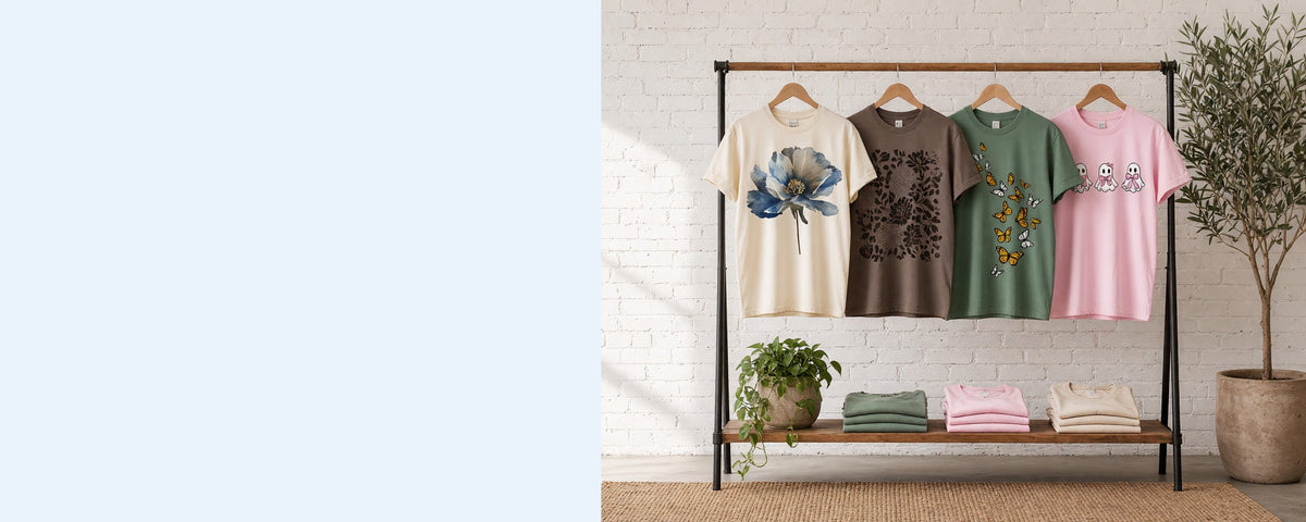 4 printed garment-dyed cotton t-shirts on wooden hangers on a wood and iron clothing rack against a sunlit white brick wall, with an olive tree, trailing pothos and folded tees on the shelf below