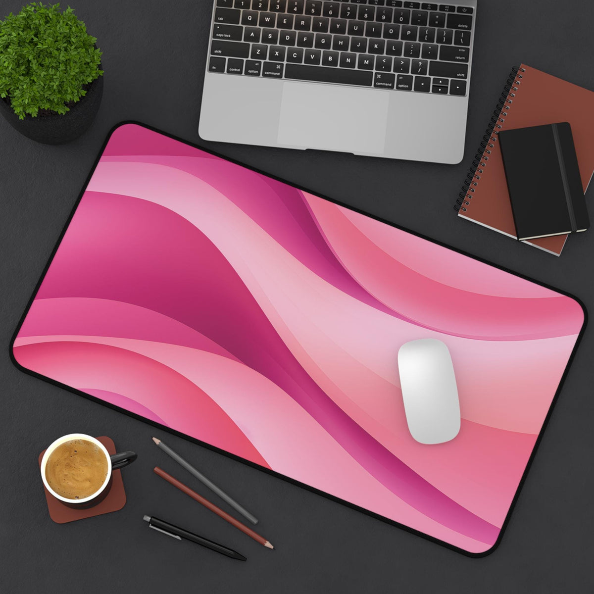 Abstract Desk Mat in pink on a dark desk with laptop, plant, notebooks, mouse, coffee and pens, overhead, 12 × 22 inches.