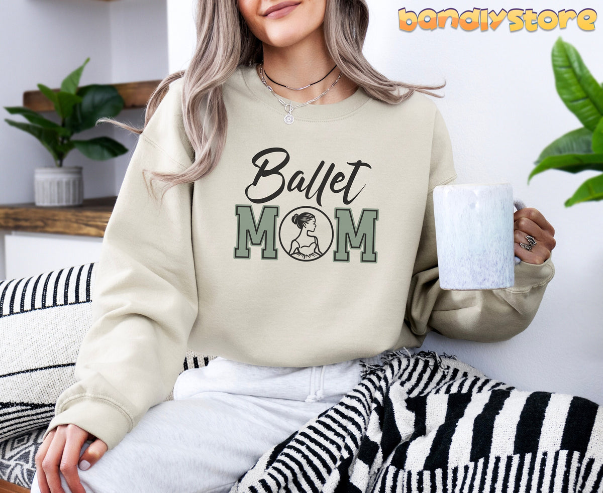 Ballet Mom Sweatshirt in sand worn by woman holding mug, seated near plants and striped blanket.