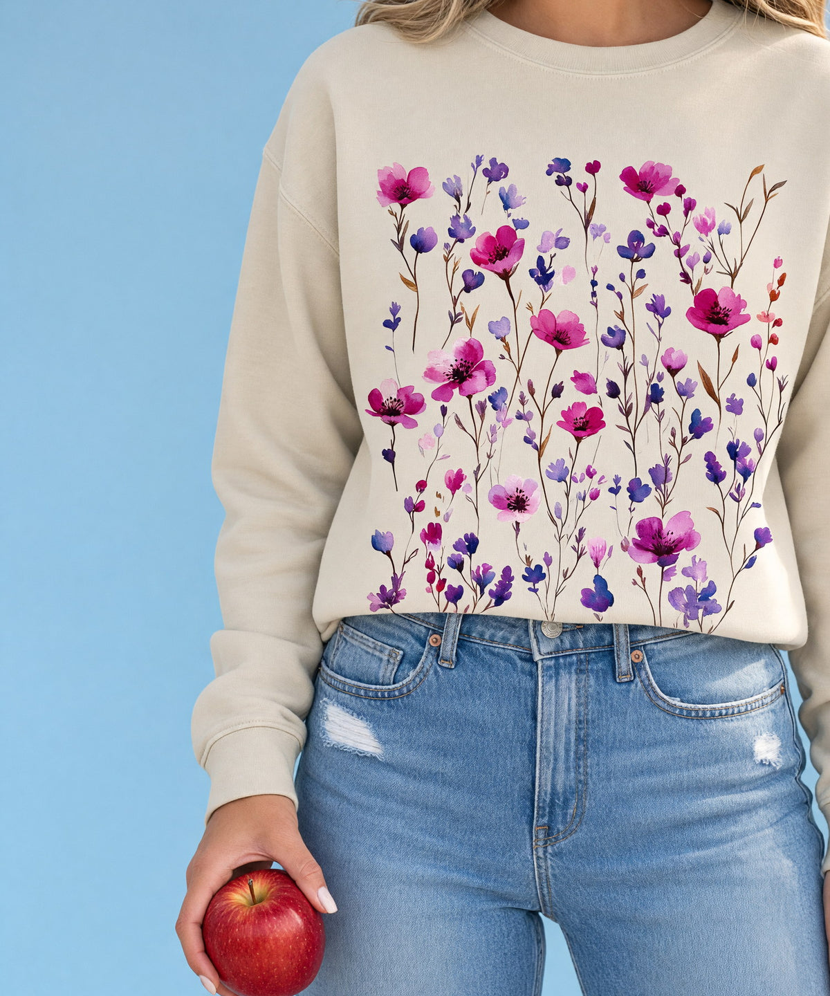 A woman in a printed crewneck sweatshirt holding an apple, cropped at the shoulders, against a sky-blue backdrop