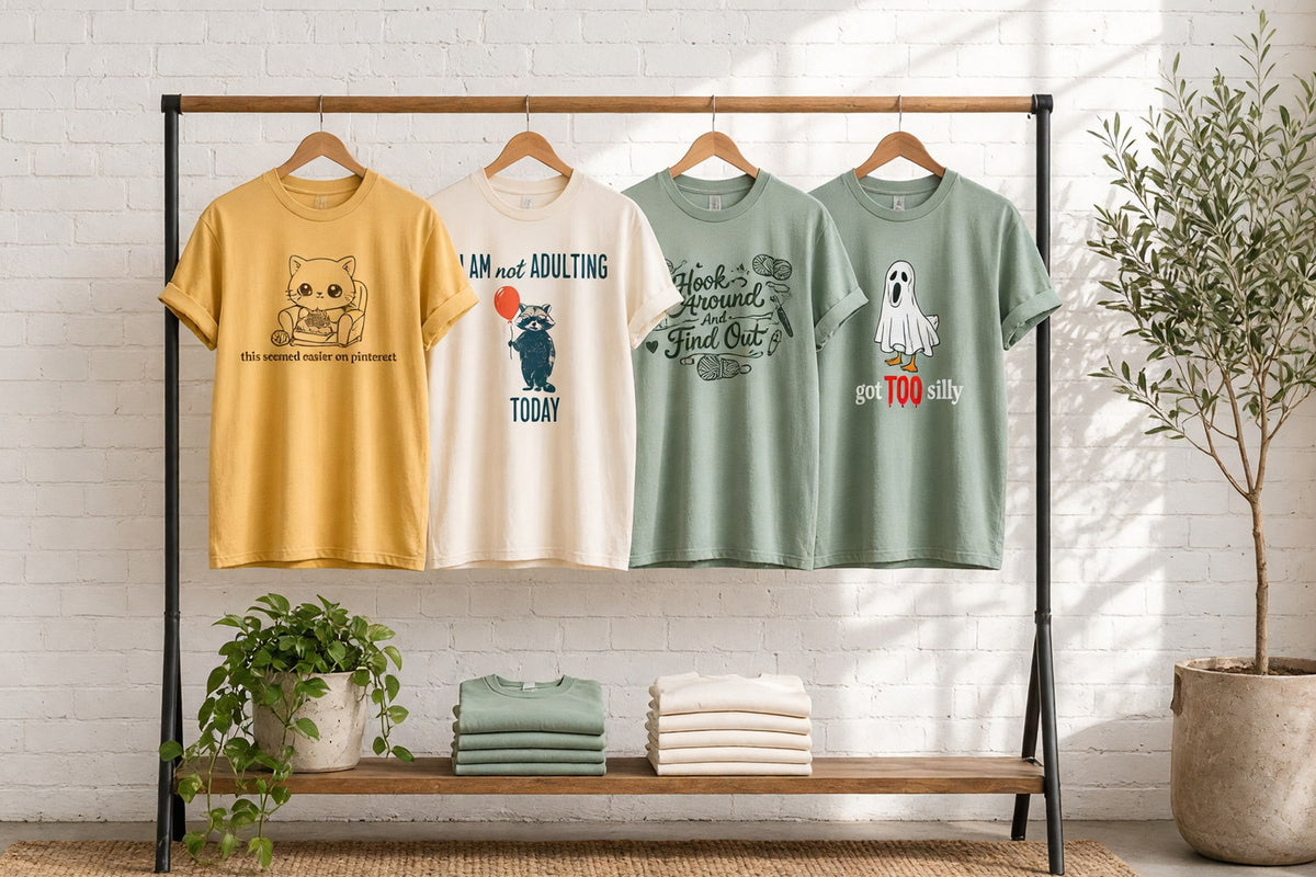 Four funny garment-dyed t-shirts with a knitting cat, a raccoon who is not adulting today, Hook Around And Find Out and a goose that got too silly on wooden hangers against a sunlit white brick wall