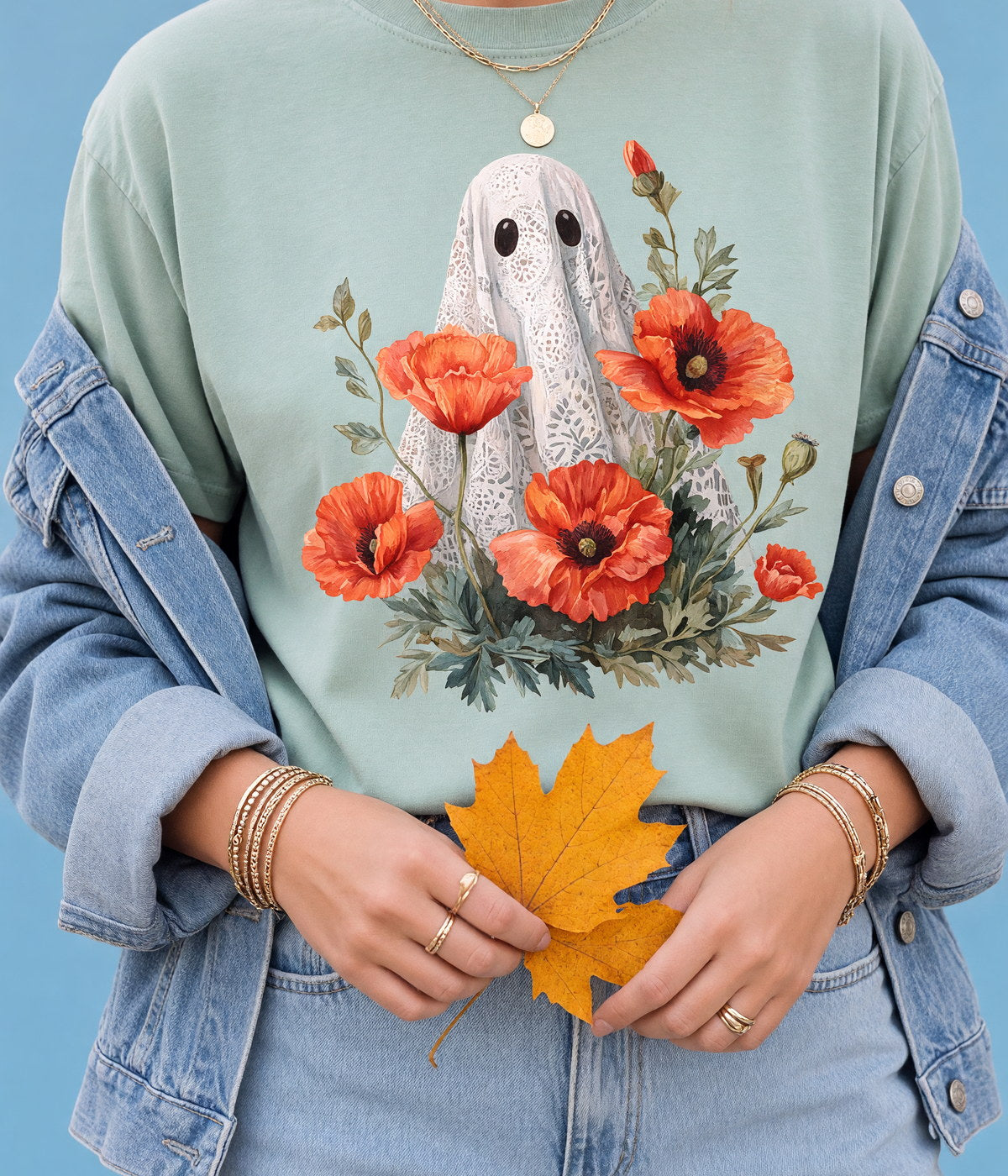 A woman in a printed t-shirt and denim jacket holding maple leaves, cropped at the shoulders, against a sky-blue backdrop