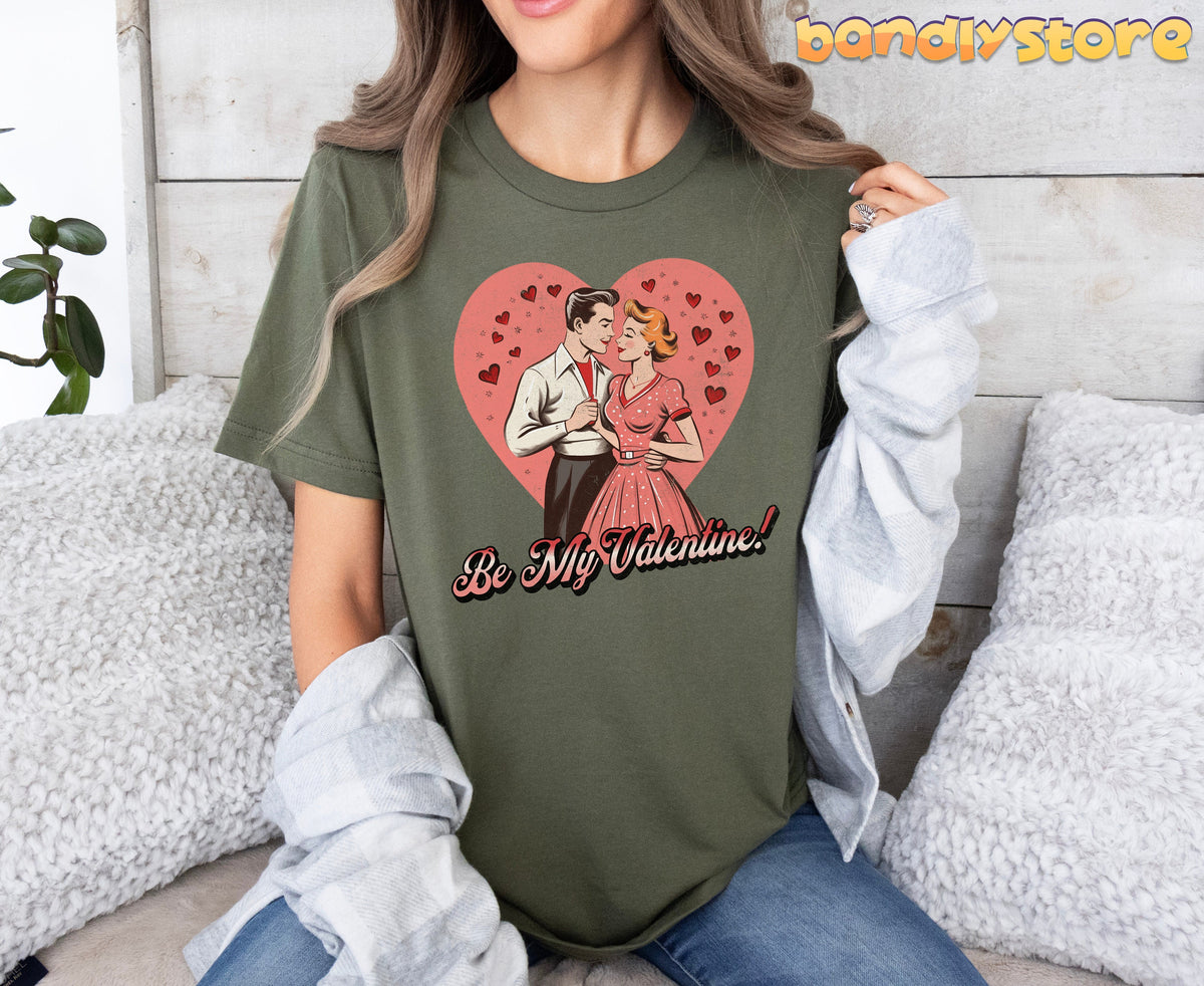 Be Mine Valentine T-Shirt in military green front view on woman seated among fluffy pillows by wooden wall.