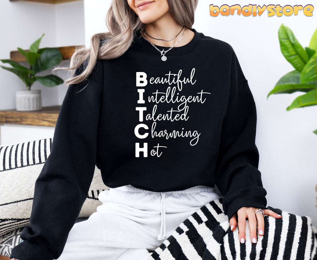 Bitch Definition Sweatshirt black on a seated woman amid plants and striped cushions.