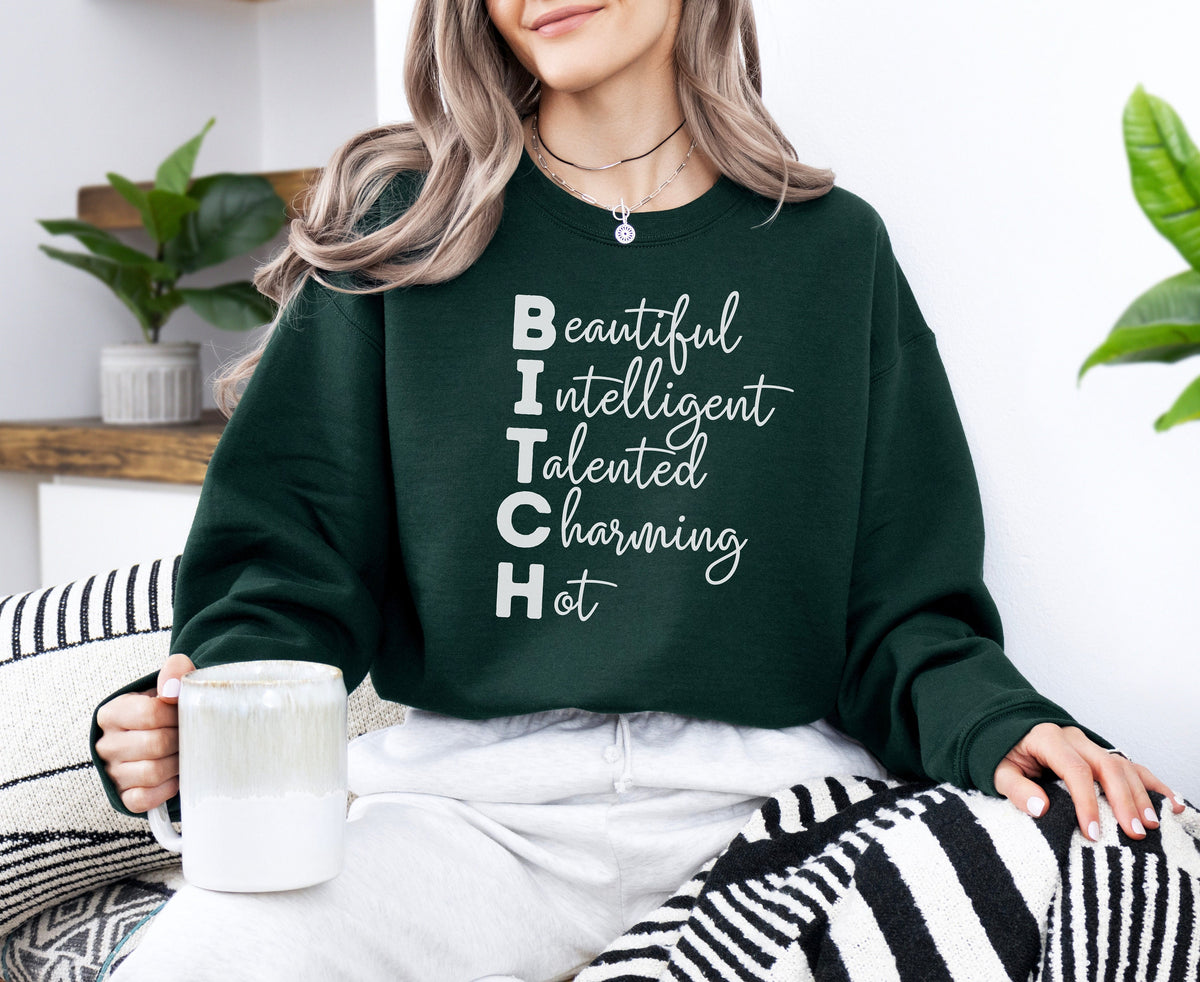 Bitch Definition Sweatshirt in forest green on seated woman holding mug by plants and striped blanket.