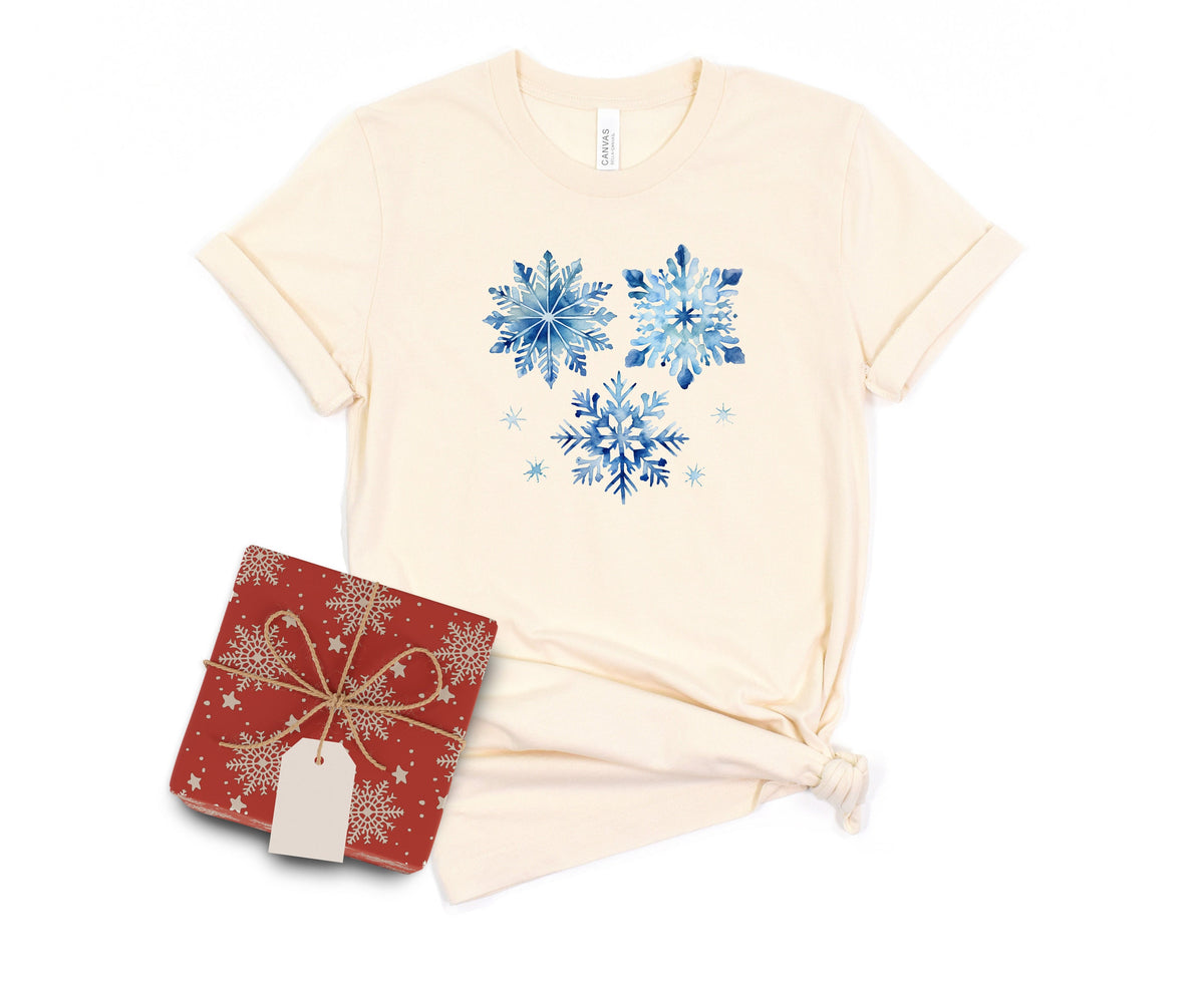 Watercolor Snowflakes T-Shirt in natural laid flat on a white background next to a red gift box.
