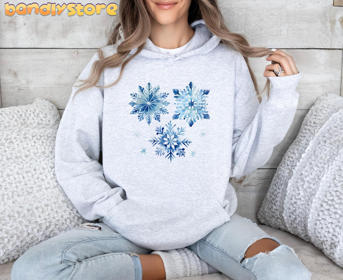 Watercolor Snowflakes Hoodie in ash, front view on a seated person against a wooden wall with white pillows.