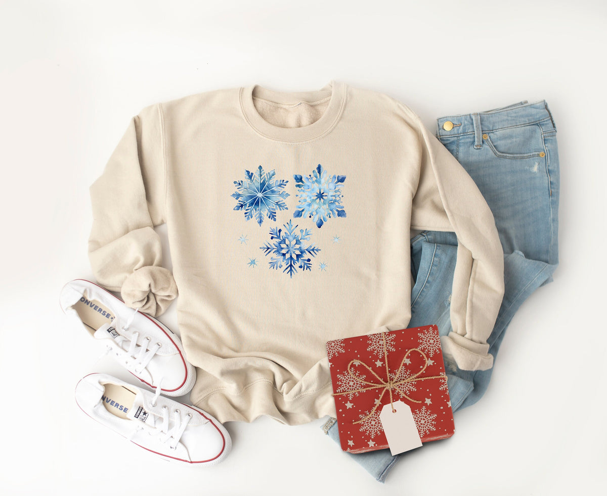 Blue Watercolor Snowflakes Sweatshirt in sand flat-laid with white sneakers, blue jeans and red gift box.