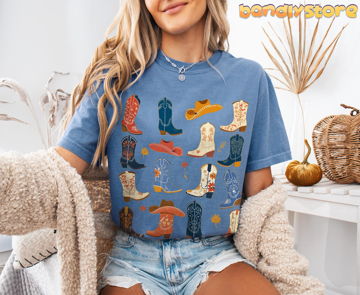 Boho Cowgirl Boots T-Shirt in Blue Jean, worn front-facing amid dried plants, a pumpkin and woven dรฉcor.
