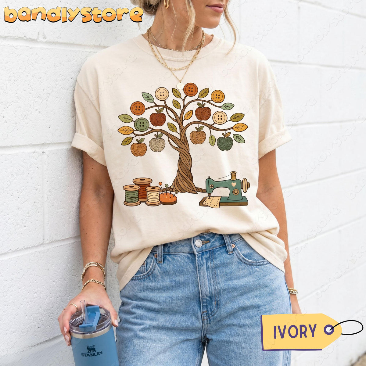 Boho Sewing T-Shirt in ivory, front view on a person leaning on a white brick wall holding a blue tumbler.