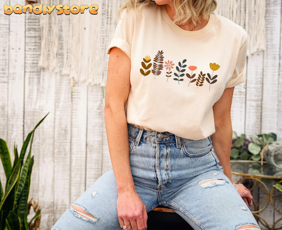 Cute Boho Wildflowers T-Shirt in soft cream, front view on seated person by white wooden wall, macrame and greenery.