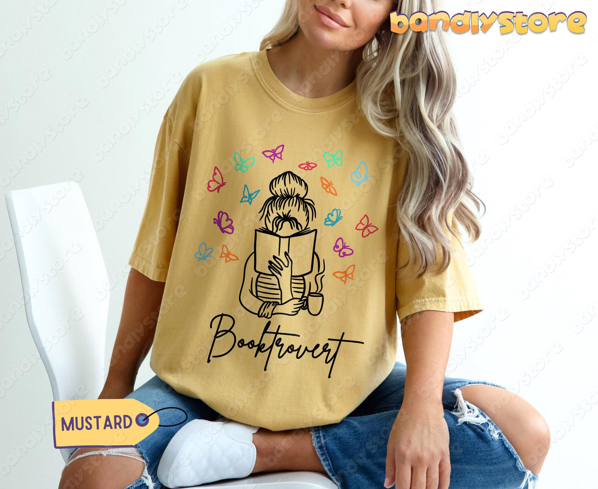 Booktrovert T-Shirt in mustard on a seated woman in ripped jeans and white sneakers beside a white chair.