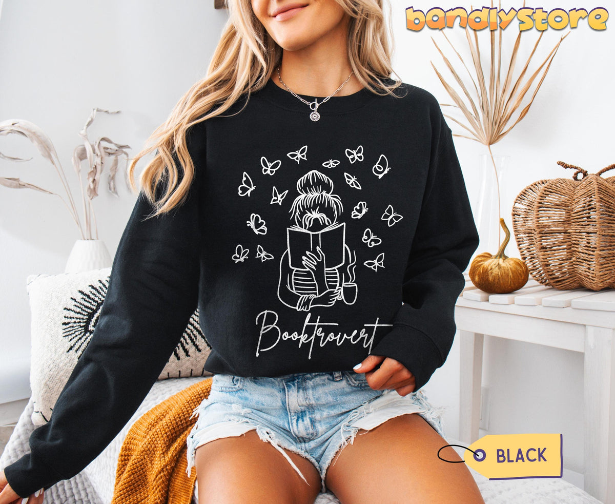 Booktrovert Sweatshirt in black, worn by a seated woman amid pillows, dried plants, a pumpkin and woven décor.