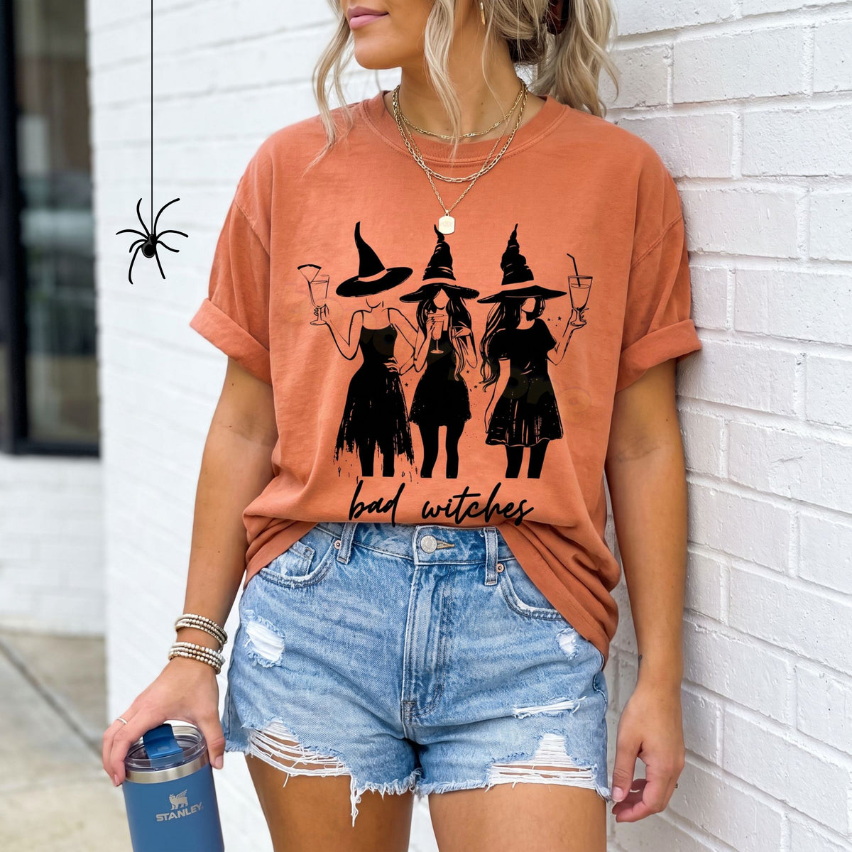 Boujee Bad Witches T-Shirt in Yam on a woman leaning against a white brick wall holding a tumbler.