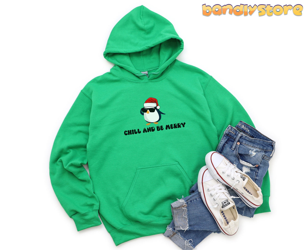 Chill And Be Merry Penguin Hoodie in Irish green laid flat beside ripped jeans and sneakers.
