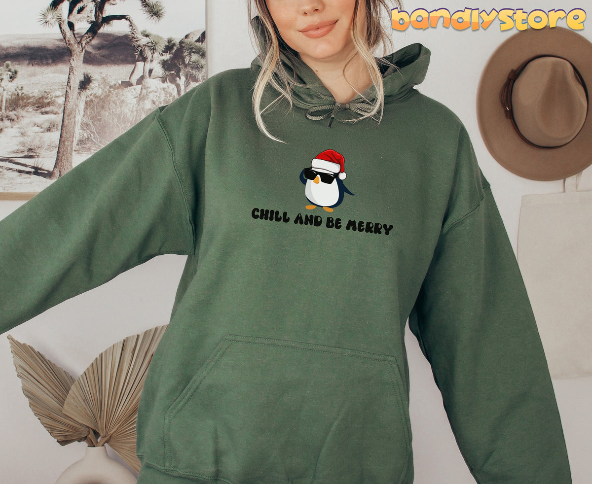 Chill And Be Merry Penguin Hoodie in Military Green on a person facing forward beside desert wall art and hat.