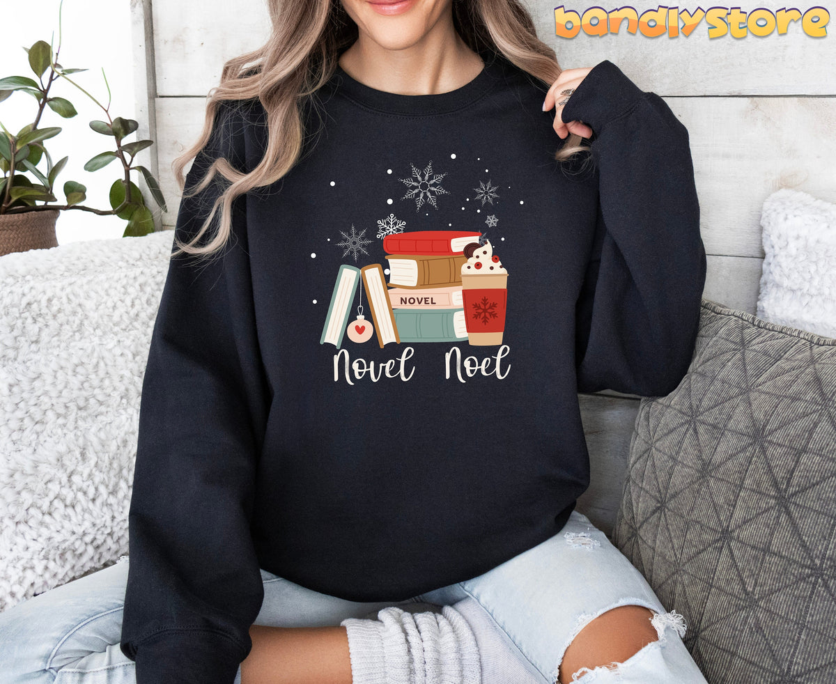 Novel Noel Christmas Sweatshirt in black worn by a woman sitting amid pillows and a plant by a wooden wall.