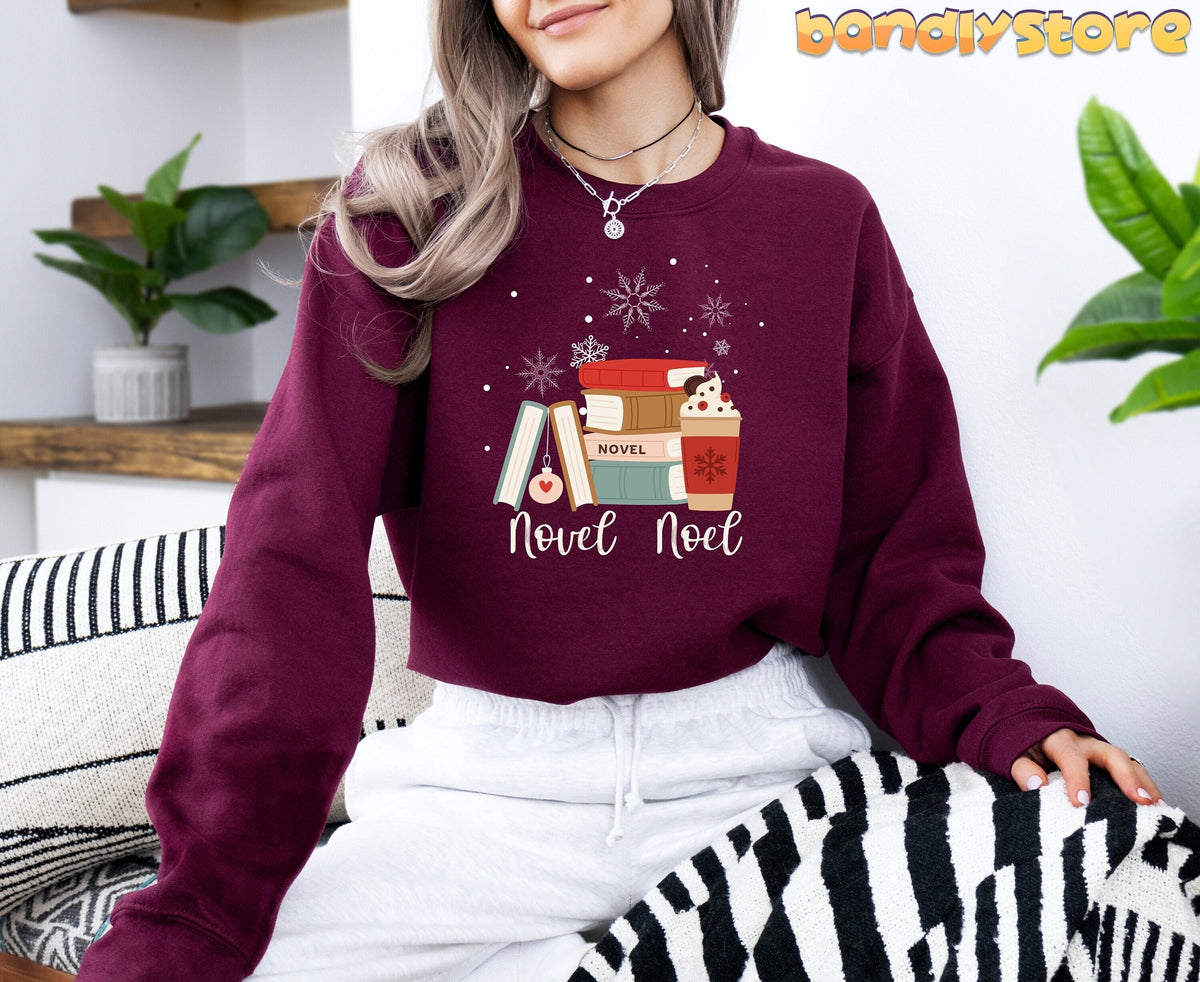 Novel Noel Christmas Sweatshirt in maroon on a woman seated indoors with plants and striped blankets.