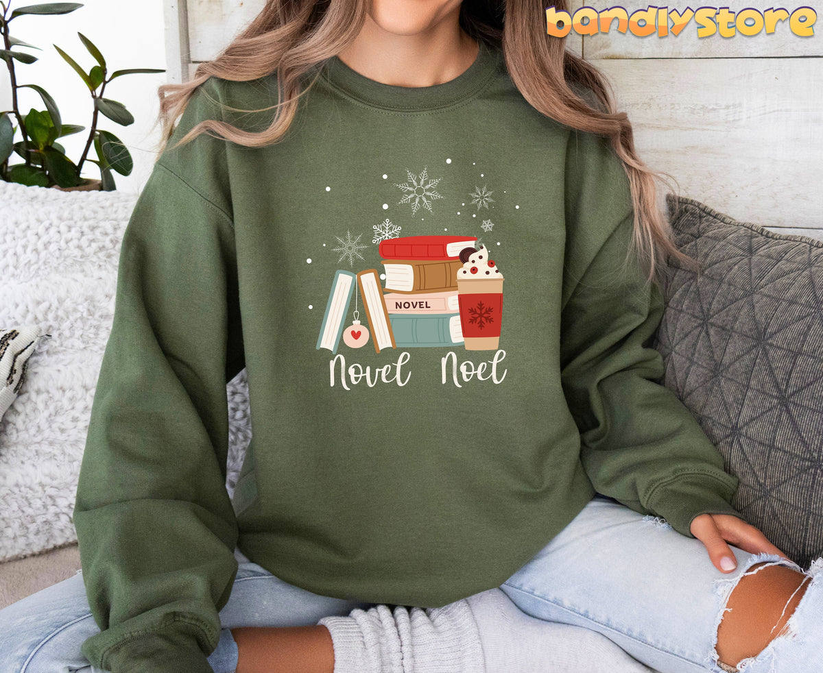 Novel Noel Christmas Sweatshirt in military green on a seated person amid pillows and a plant.