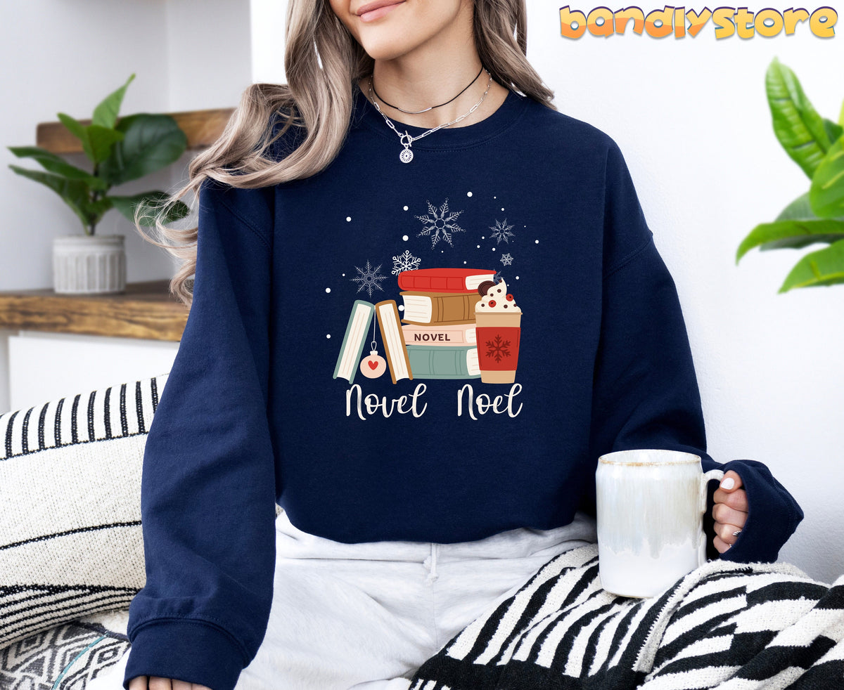Novel Noel Christmas Sweatshirt in navy worn by a seated person holding a mug amid plants and striped blankets.