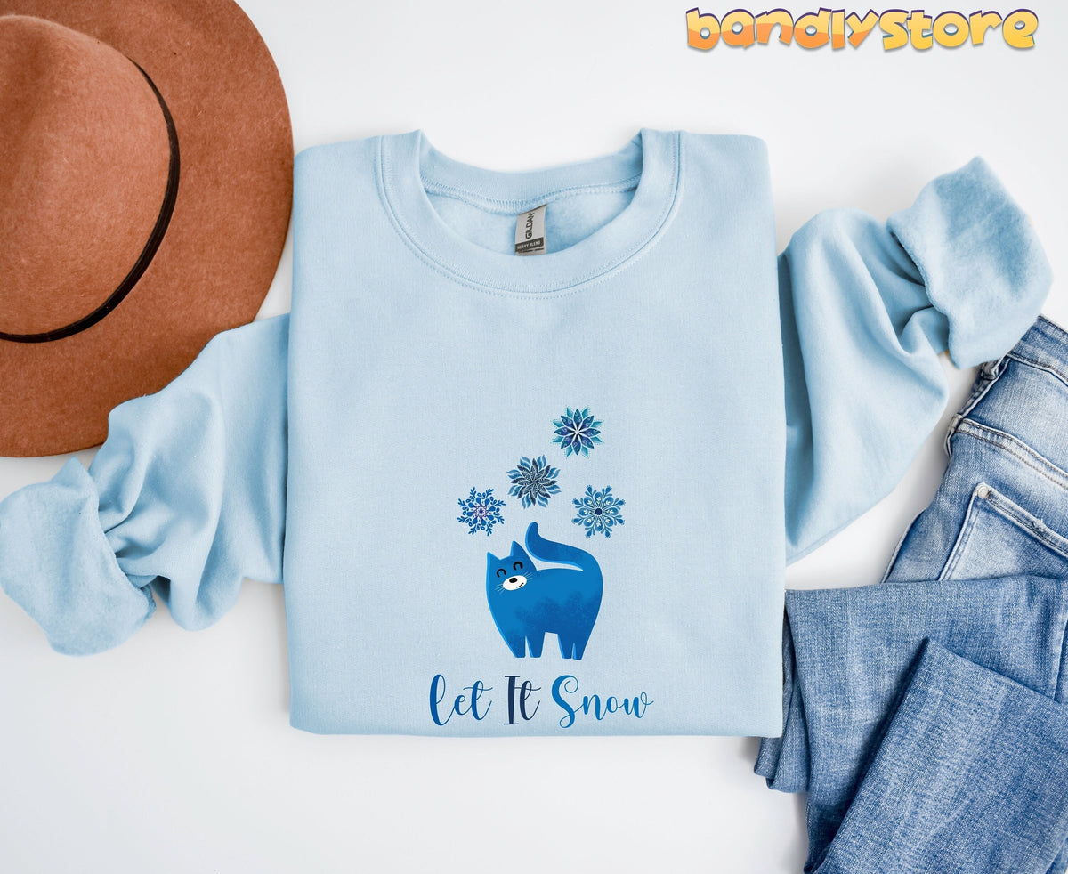 Christmas Cat Let It Snow Sweatshirt light blue folded flat beside brown hat and jeans.