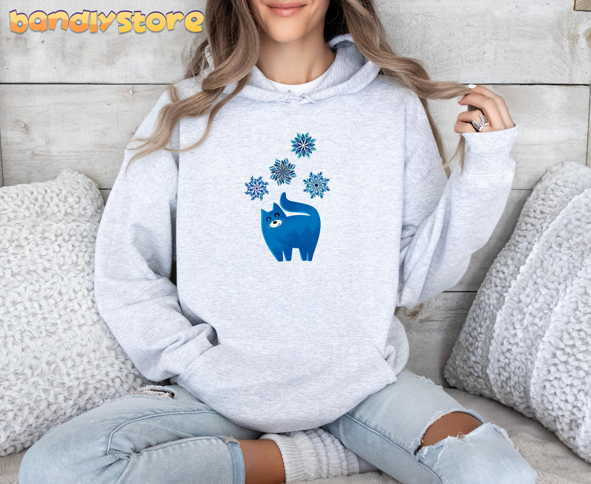 Christmas Cat And Snowflakes Hoodie in ash, front view on a seated model amid white pillows and wood wall.