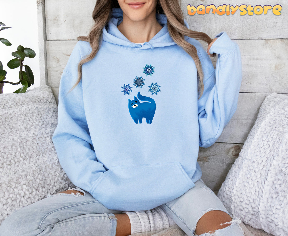 Christmas Cat And Snowflakes Hoodie in light blue worn by a seated person against a wooden wall with pillows and a plant.