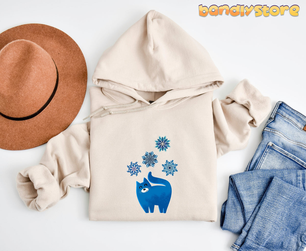 Christmas Cat And Snowflakes Hoodie in Sand, flat lay with brown hat left and blue jeans right on white background.