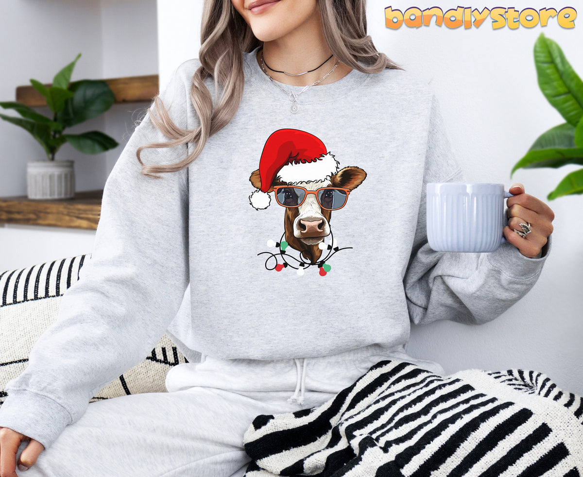 Christmas Cow Sweatshirt in ash worn by a seated person holding a mug beside plants and striped textiles.