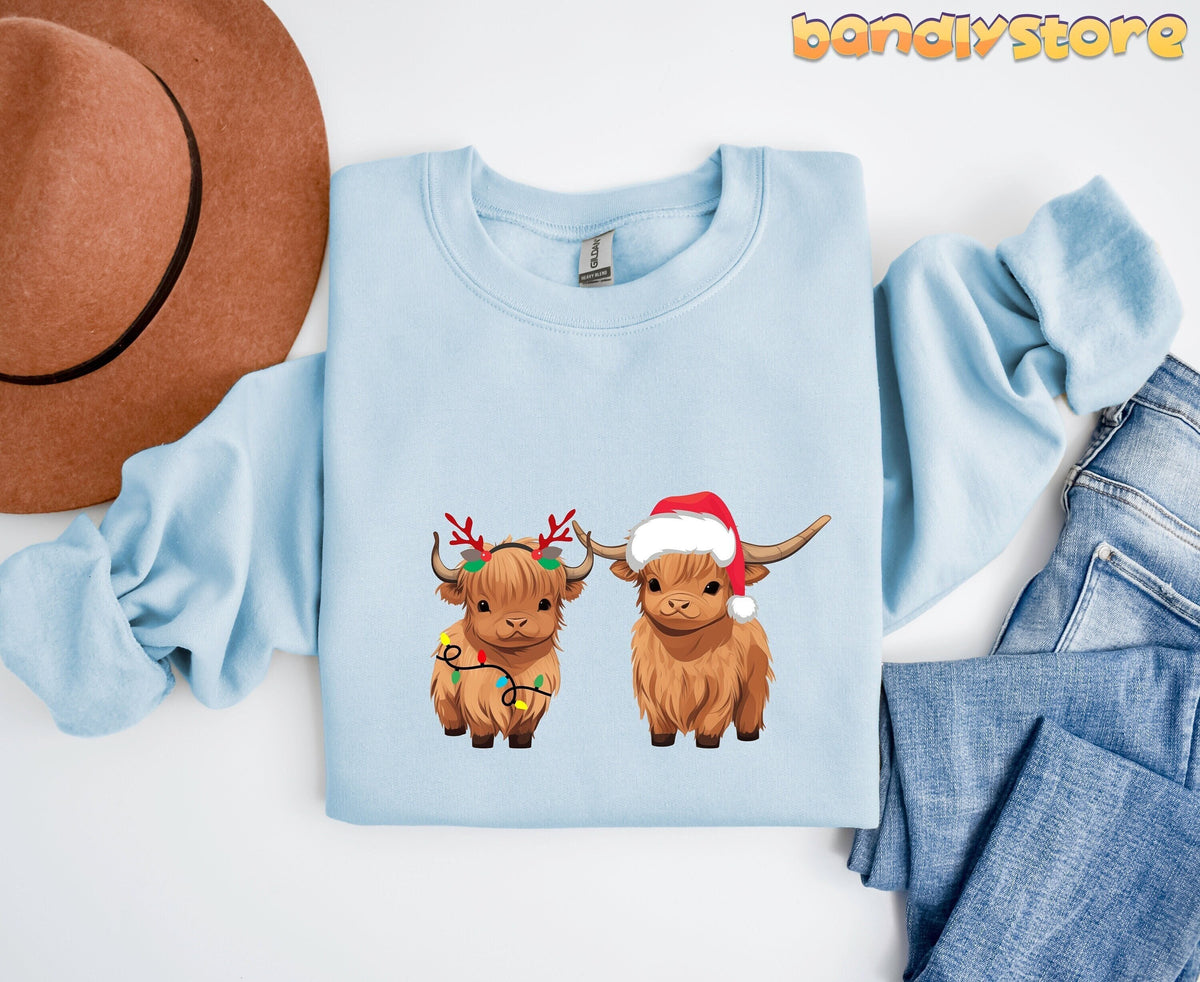 Christmas Highland Cow Sweatshirt in light blue laid flat from above beside a brown hat and blue jeans.