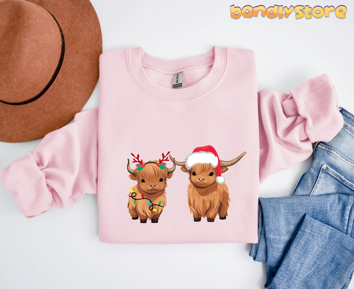 Christmas Highland Cow Sweatshirt in light pink flat lay beside brown hat and blue jeans.