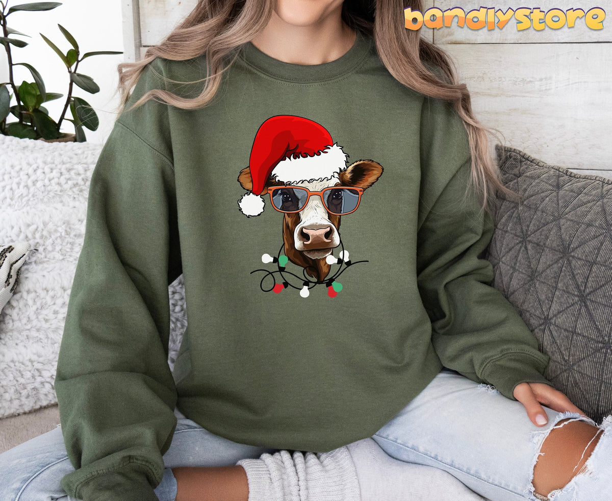 Christmas Cow Sweatshirt in military green, worn front-facing by a seated person amid plants, pillows and wood wall.