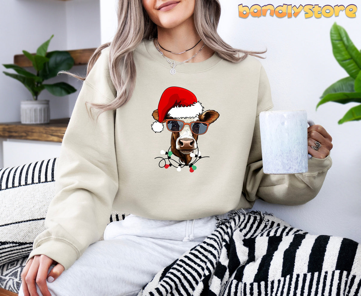 Christmas Cow Sweatshirt in sand on a seated person holding a mug beside plants and pillows.