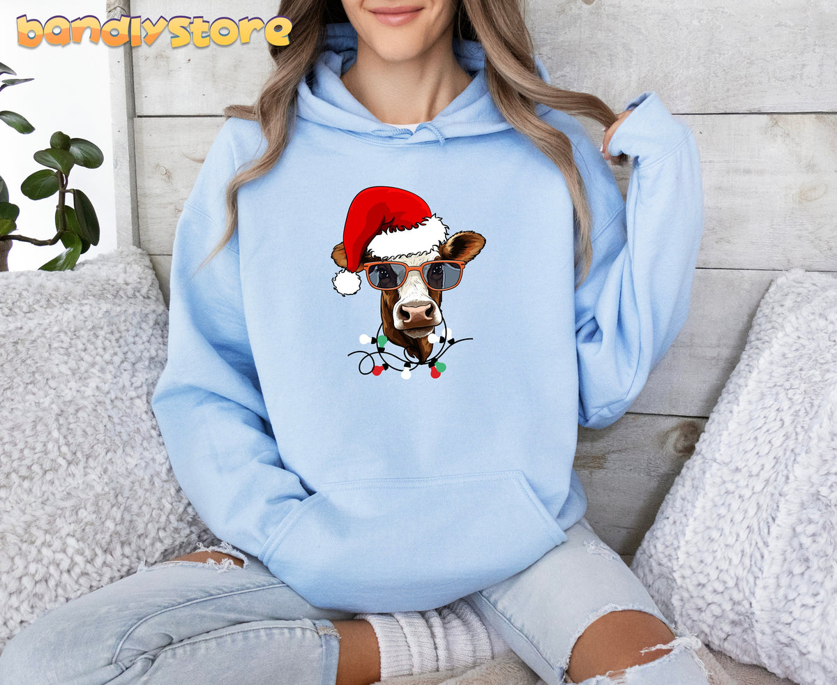 Christmas Highland Cow Hoodie in light blue on a seated person by pillows, plant and wood wall.