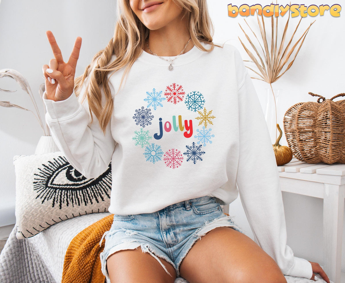 Jolly Snowflake Sweatshirt in white, front view on seated person with pillows and dried plants nearby.