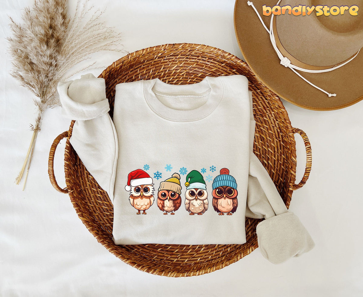 Cute Christmas Owl Sweatshirt in sand folded in a wicker basket beside pampas grass and a brown hat.