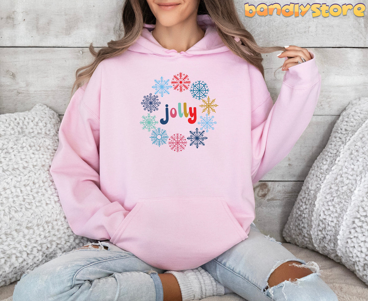 Christmas Snowflake Jolly Hoodie in light pink on seated person amid white pillows against wooden wall.