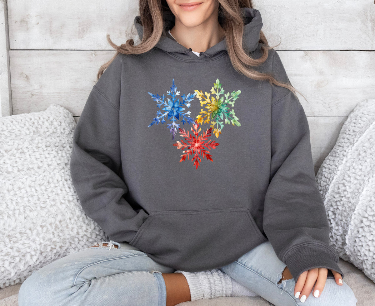 Colorful Watercolor Snowflakes Hoodie in charcoal, modeled front-on while seated amid white pillows on a couch.