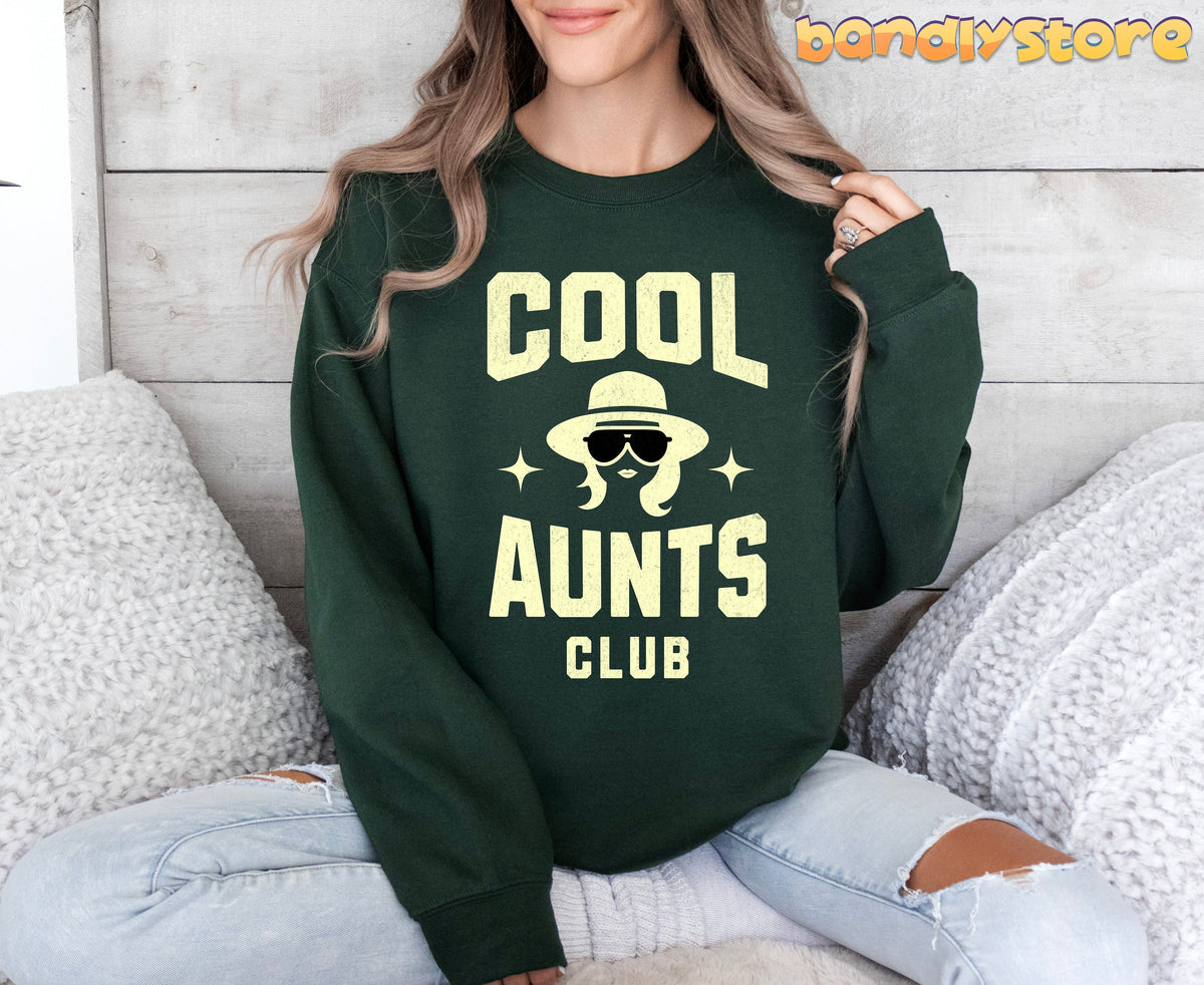 Cool Aunts Club Sweatshirt in forest green on a seated person against a wooden wall with white fluffy pillows.