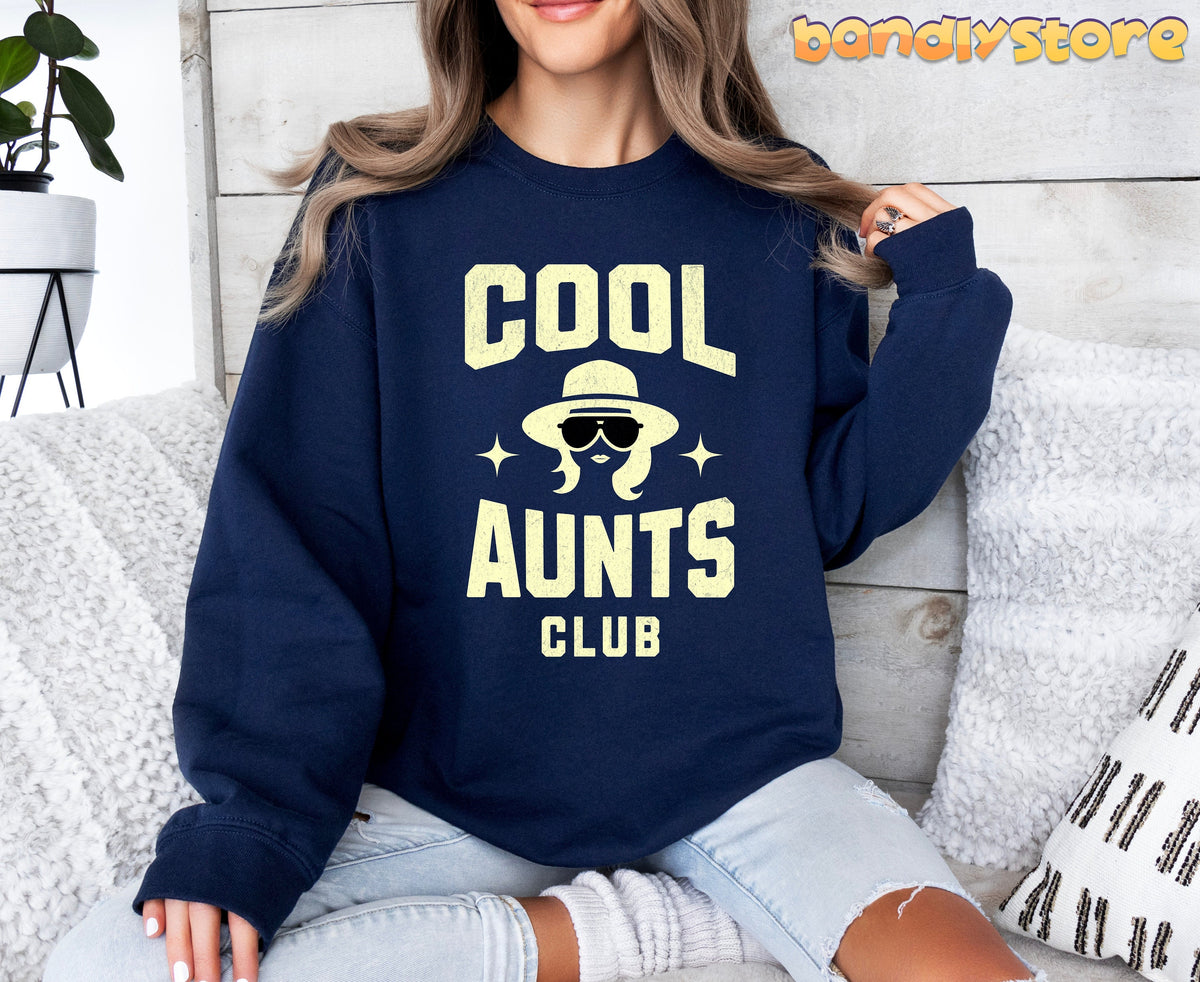 Cool Aunts Club Sweatshirt in navy worn by a seated woman among white pillows and a plant against a wooden wall.
