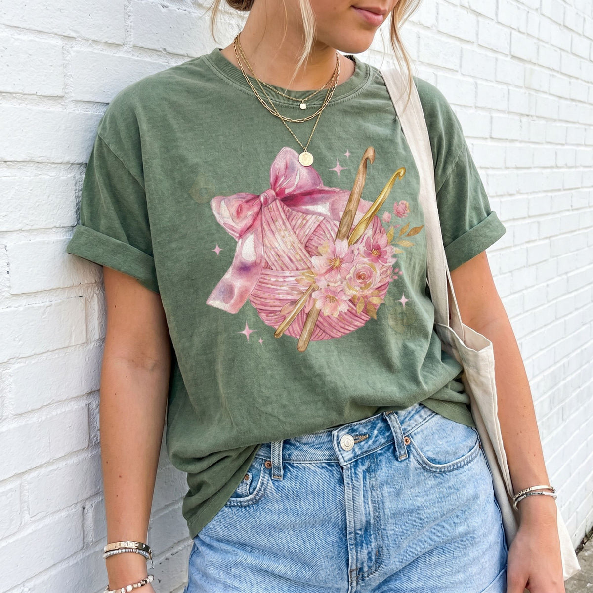 Coquette Crocheting T-Shirt in moss on a person against a white brick wall with jeans and tote bag.