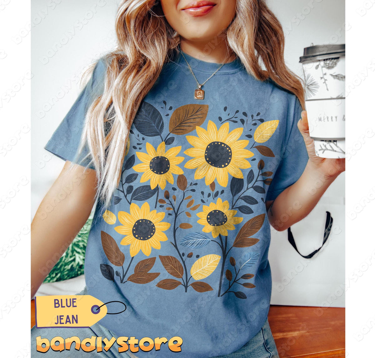 Cottagecore Sunflower T-Shirt in Blue Jean on a person holding a cup, front torso view indoors by wood and bag.