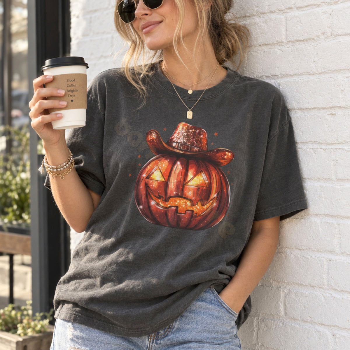 Cowboy Jack-O-Lantern T-Shirt in Pepper, worn by a woman outdoors holding coffee against a white brick wall.